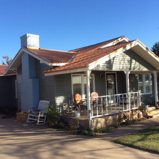 A light green house with a porch and a red roof, a chimney, and a blue sky in the background.