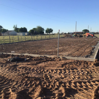 Construction site, dirt, tractor tracks, concrete foundation, fenced field, backhoe, clear sky.