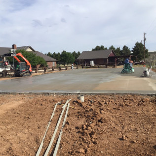 Construction site with freshly poured concrete. Workers use machinery in front of buildings, brown dirt foreground.