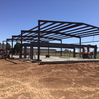 Steel frame of a building under construction on a dirt lot. Blue sky. Construction workers visible.