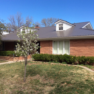Brick house with a gray metal roof and dormers, surrounded by grass, bushes, and a blooming tree, under a blue sky.