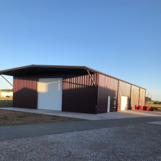 Maroon metal warehouse with white doors and a long driveway under a blue sky.