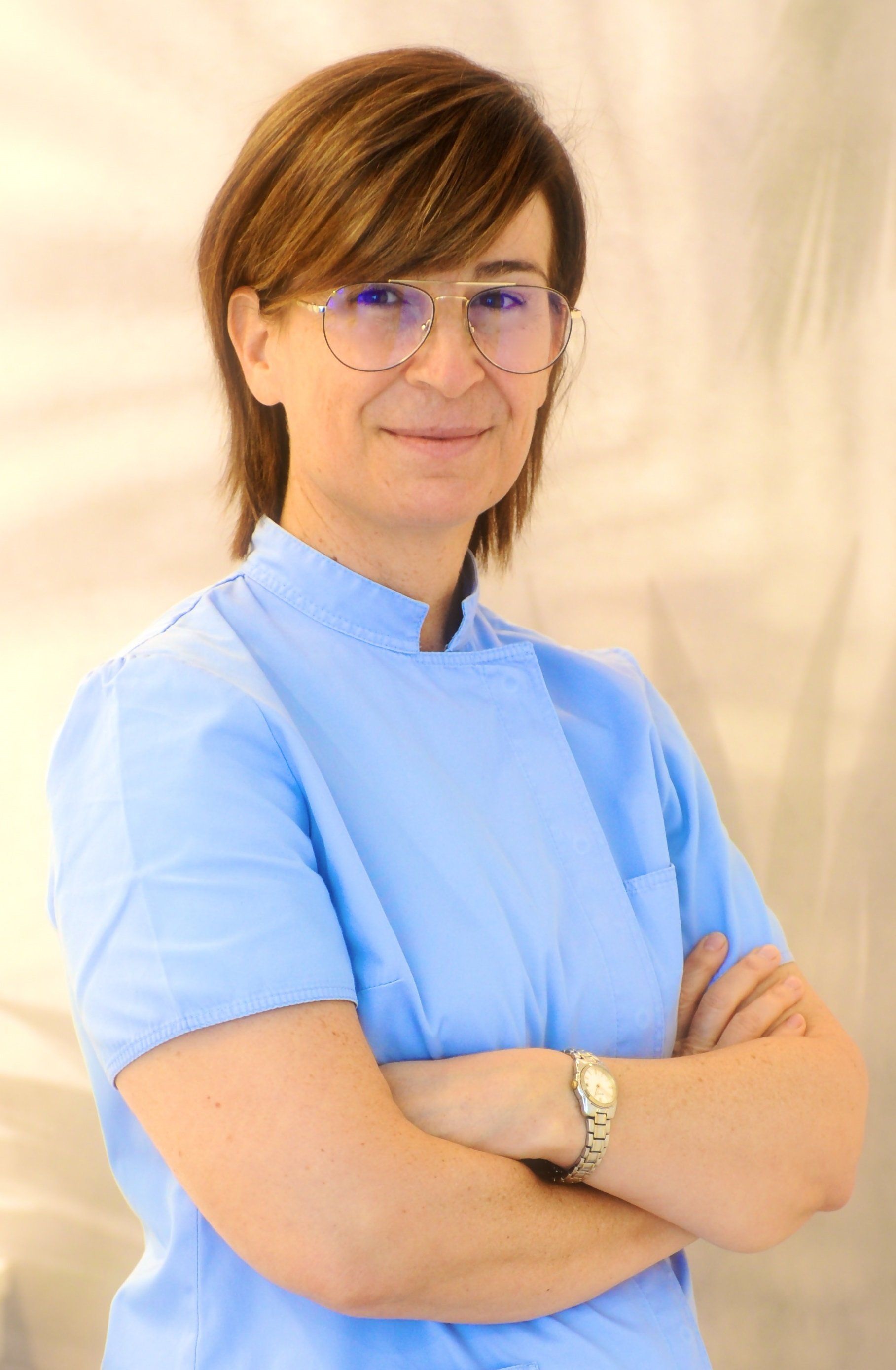 Una donna con un camice blu e gli occhiali è in piedi con le braccia incrociate.