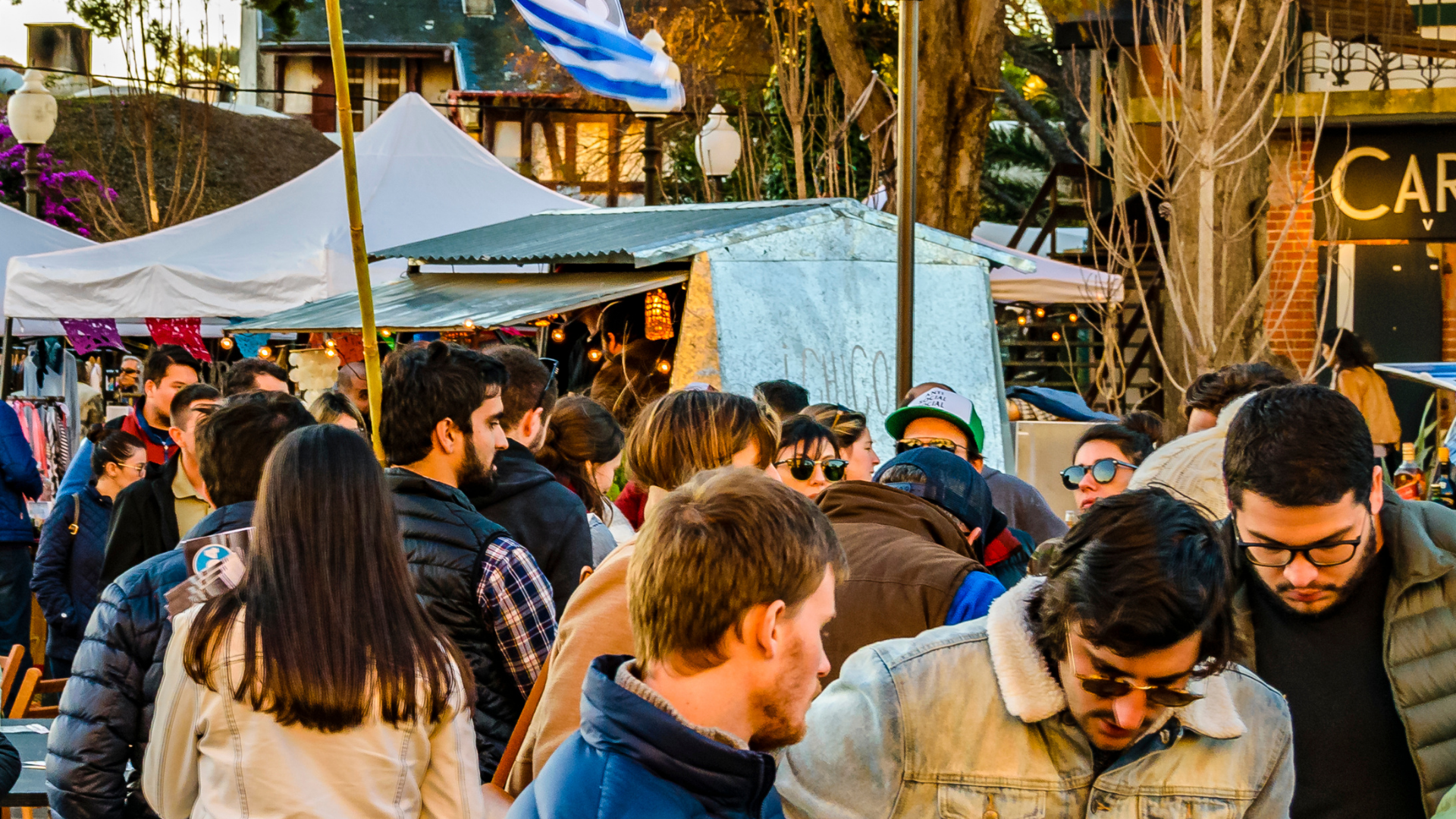 Crowded outdoor market with people browsing under white tents and blue tarps