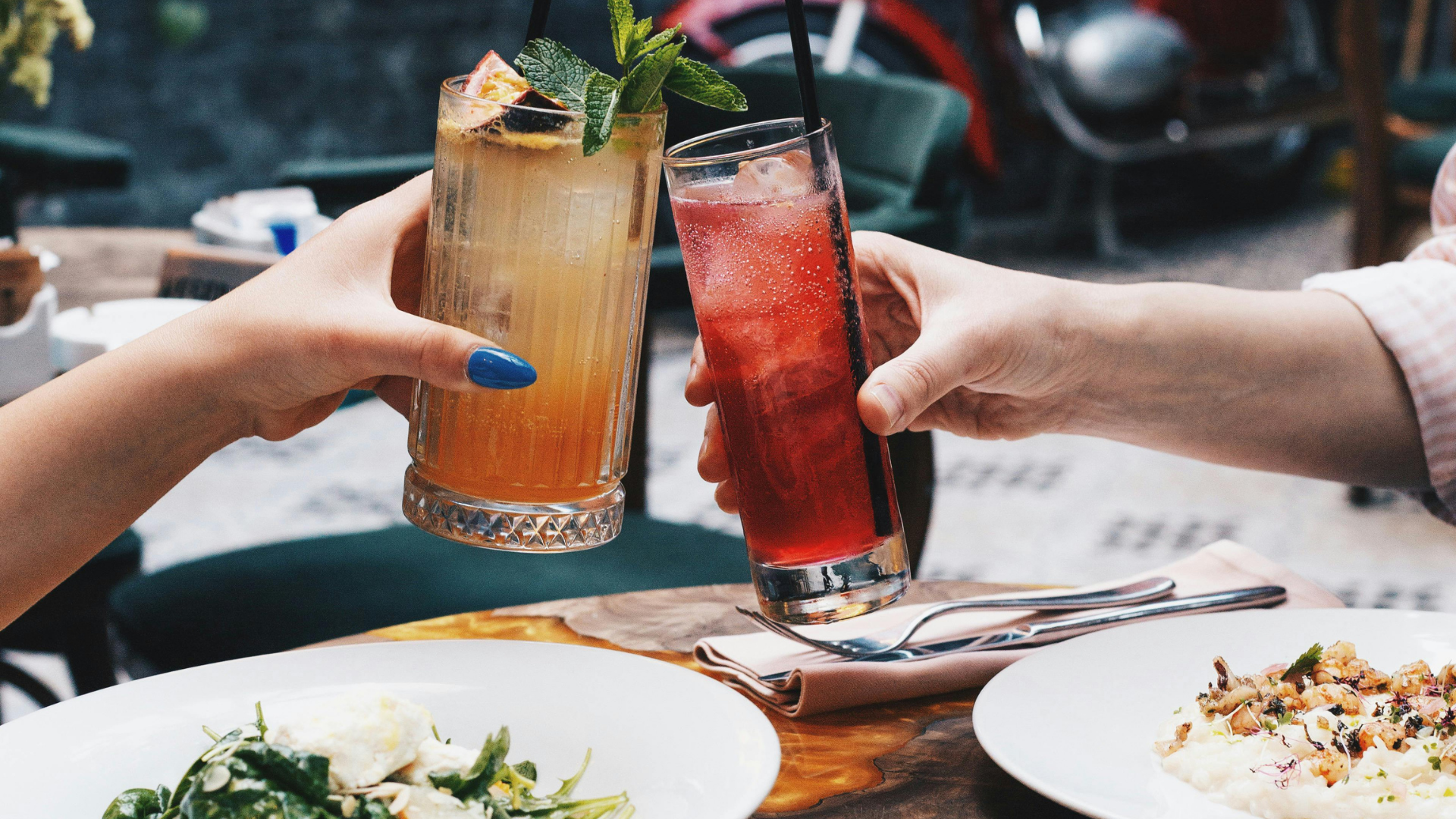 Two hands toasting with colorful cocktails over a table with plates of food outdoors
