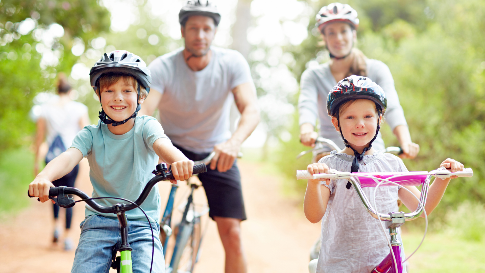Two children riding bikes on a sunny path while adults follow behind them