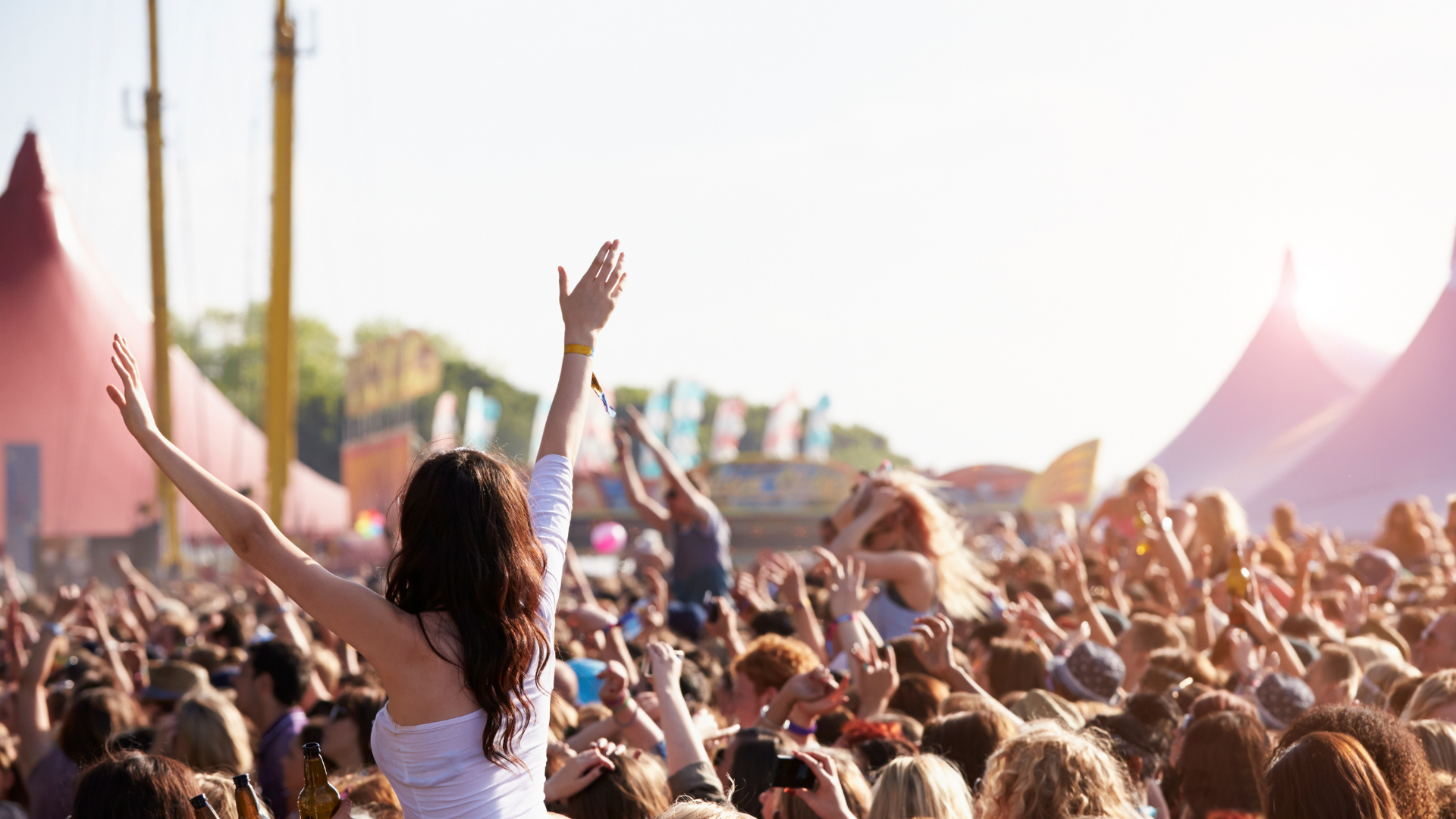Crowd cheering at an outdoor concert, with a woman raising her arms in the foreground under bright sunlight