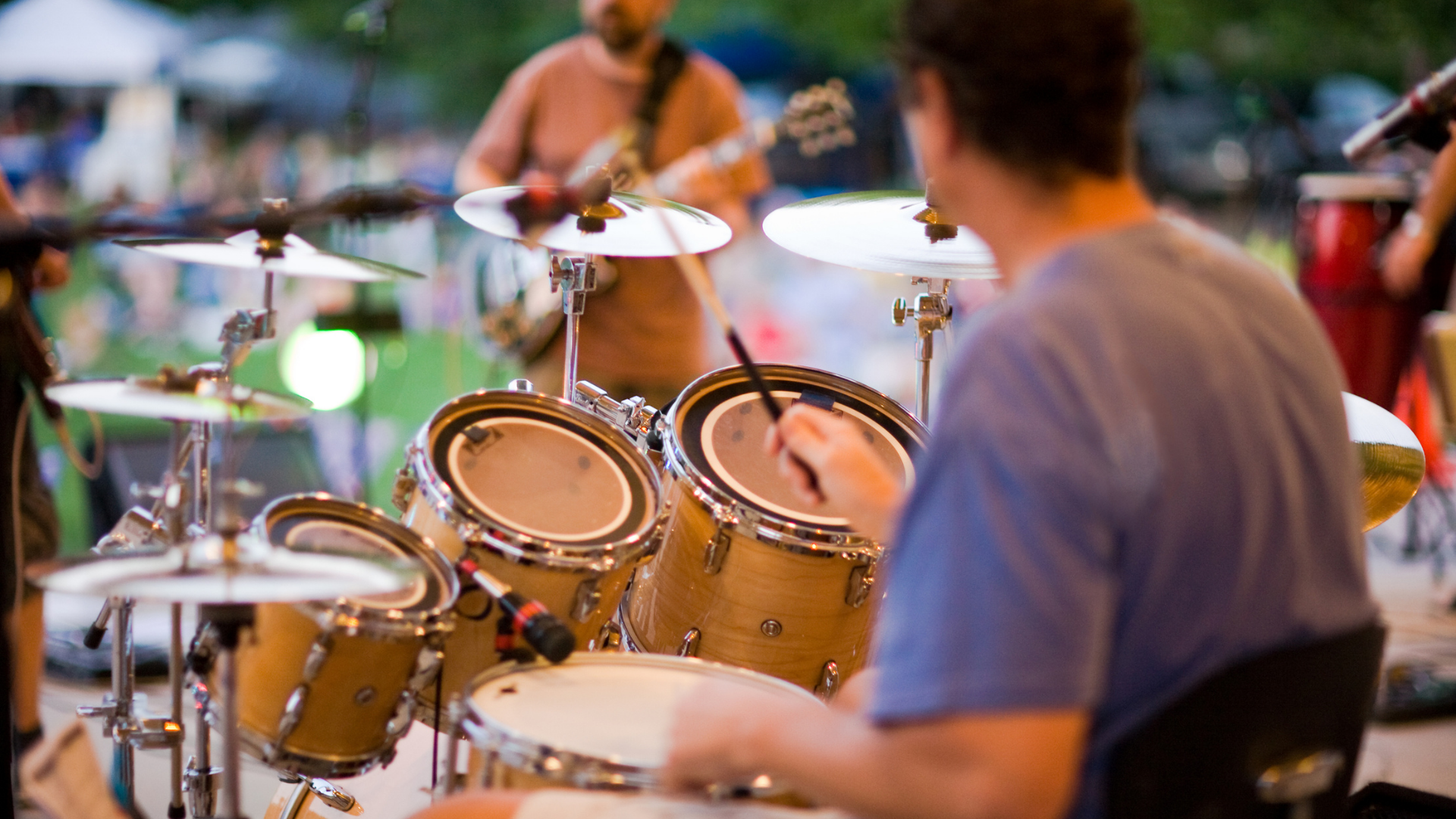 Drummer performing onstage with a tan drum kit, seen from behind in a lively outdoor setting