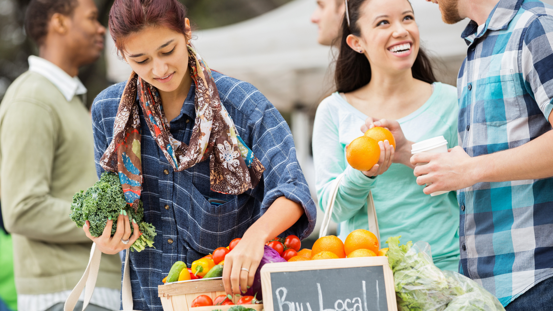 People shopping at an outdoor market, holding fresh produce beside a “Buy Local” sign