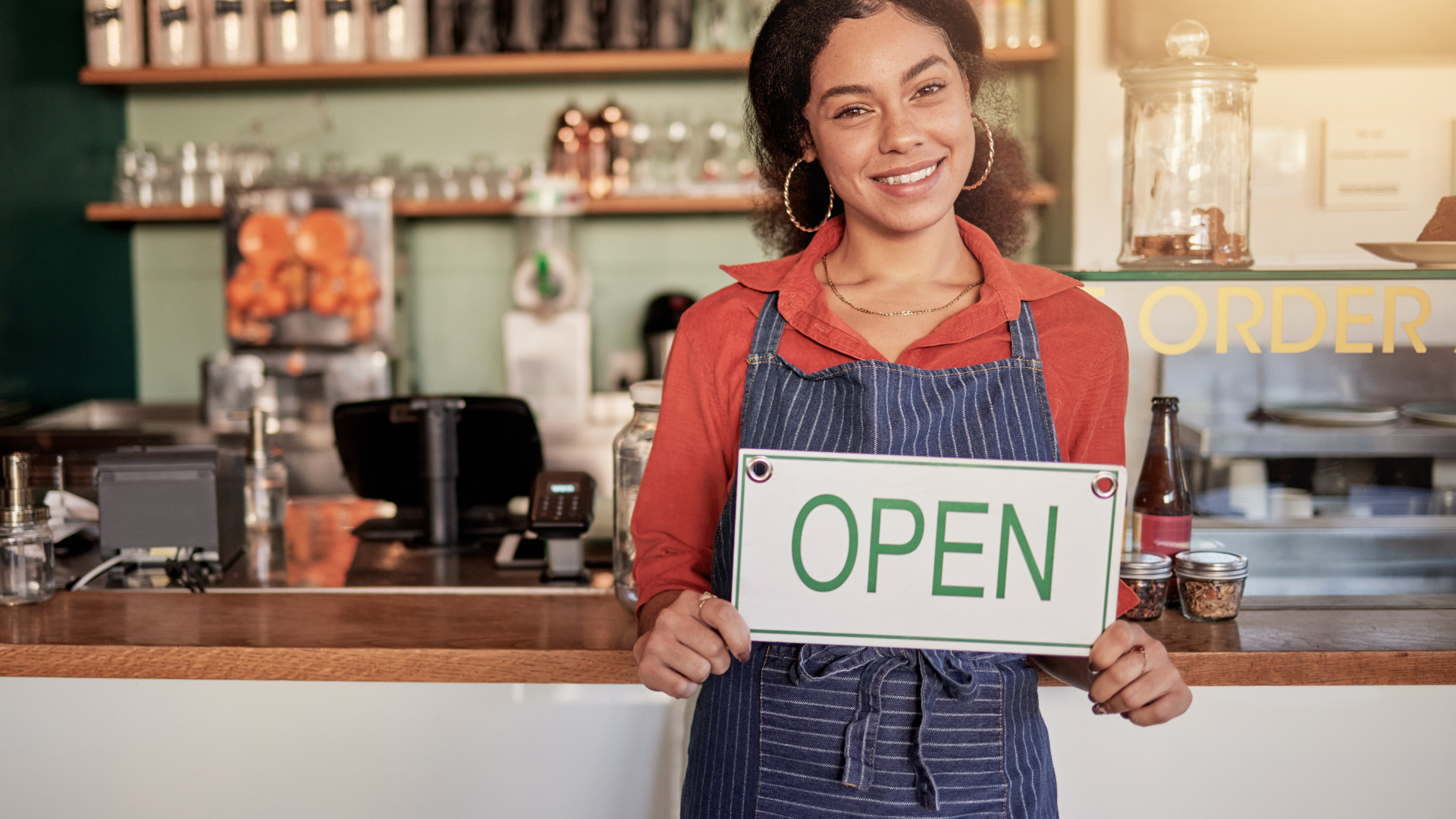 Smiling shopkeeper holding an OPEN sign at a café counter