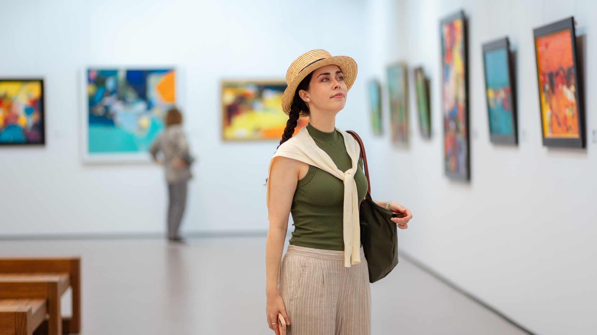 Woman in a straw hat stands in an art gallery, with colorful paintings on white walls.