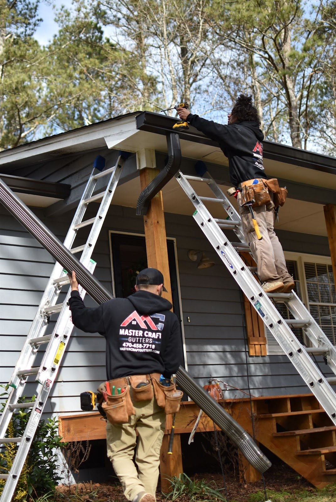 Workers on ladders repairing a house roof and gutter under a tree line.