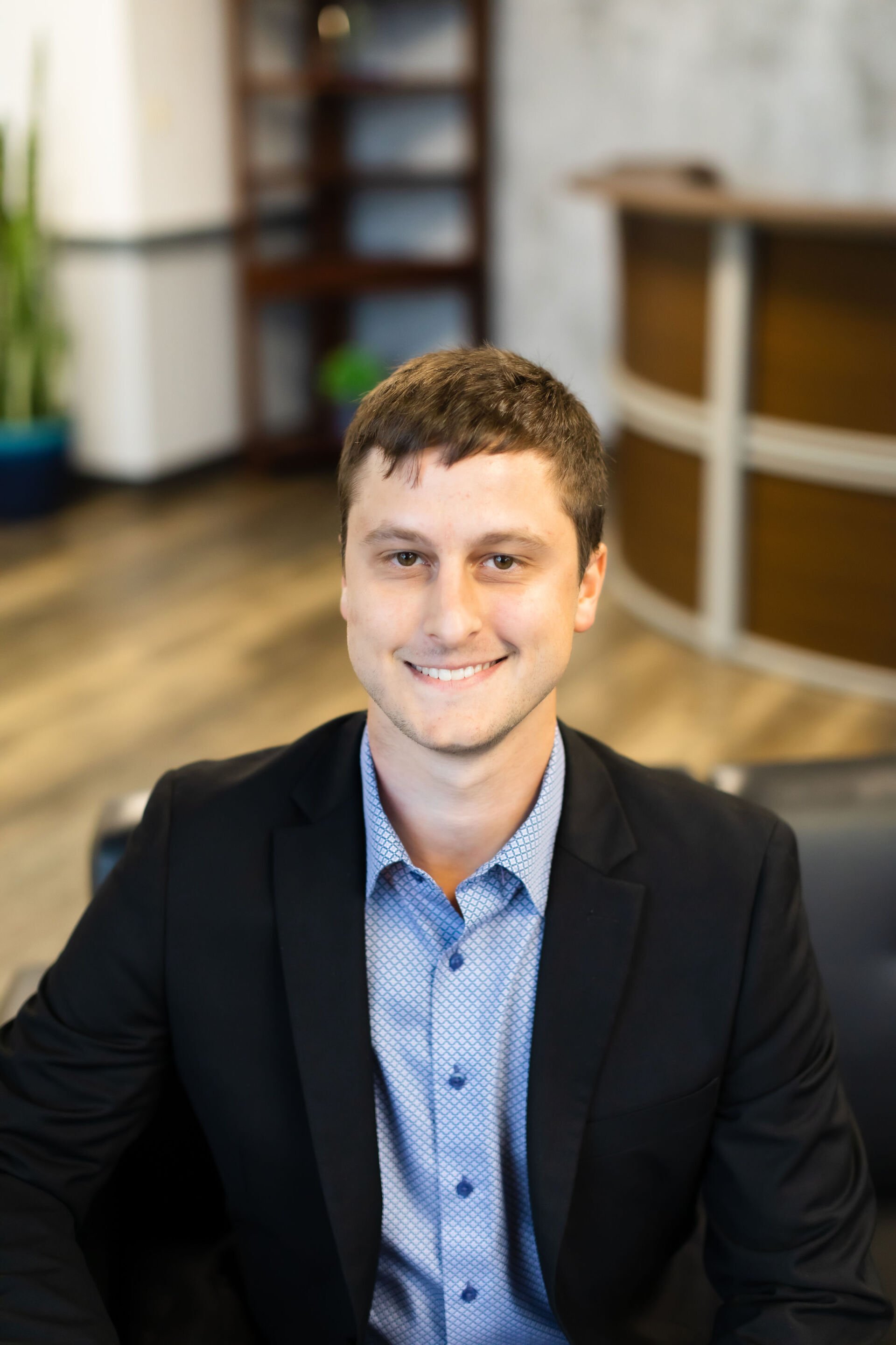 A man in a suit and blue shirt is sitting in a chair and smiling for the camera.