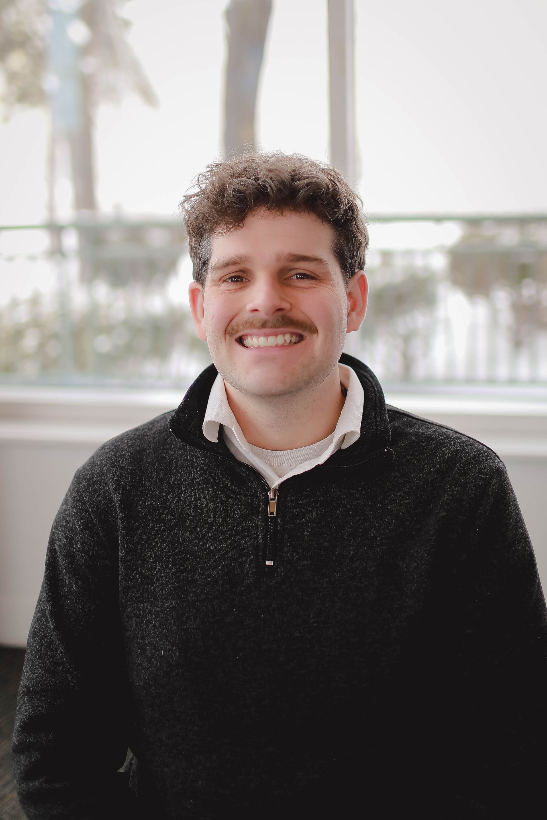 A young man with a mustache is wearing a black sweater and smiling for the camera.