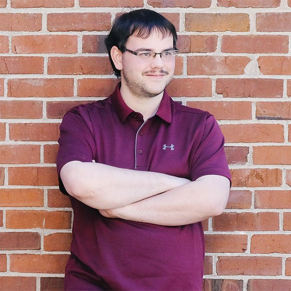 A man wearing a maroon under armour shirt is standing in front of a brick wall