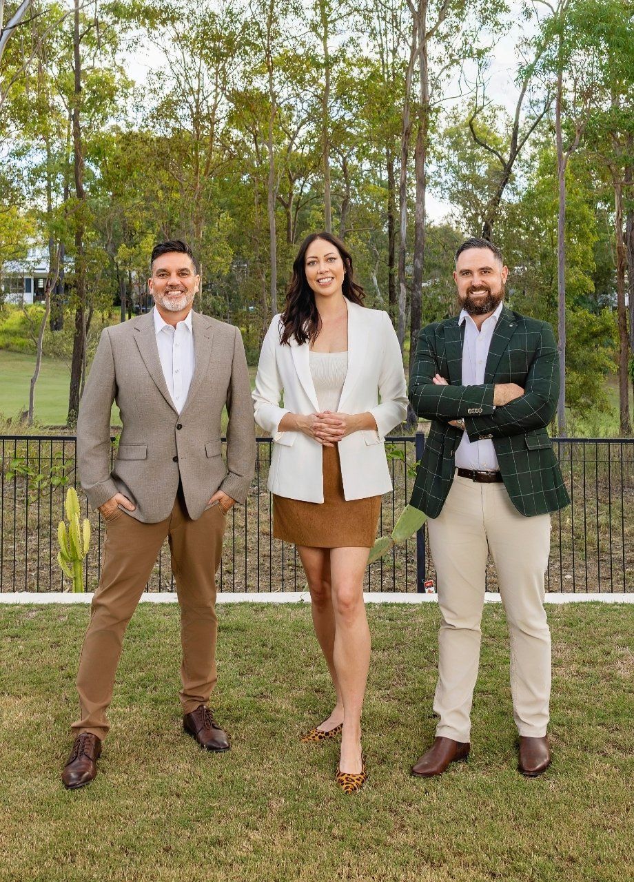 Three people posing outdoors on grass in front of trees, dressed in business casual attire.