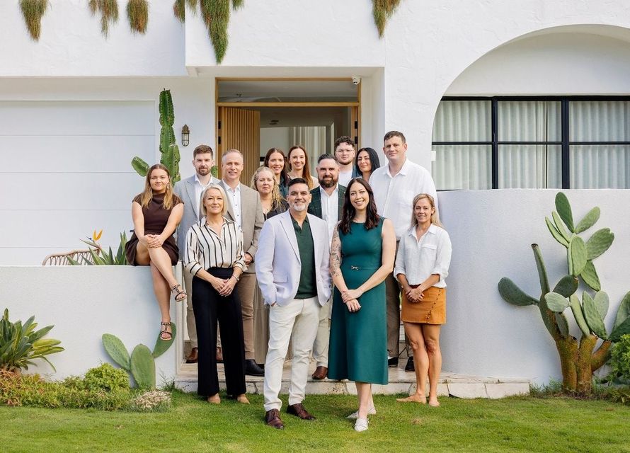 Group of people posing in front of a modern house with white walls and desert plants.