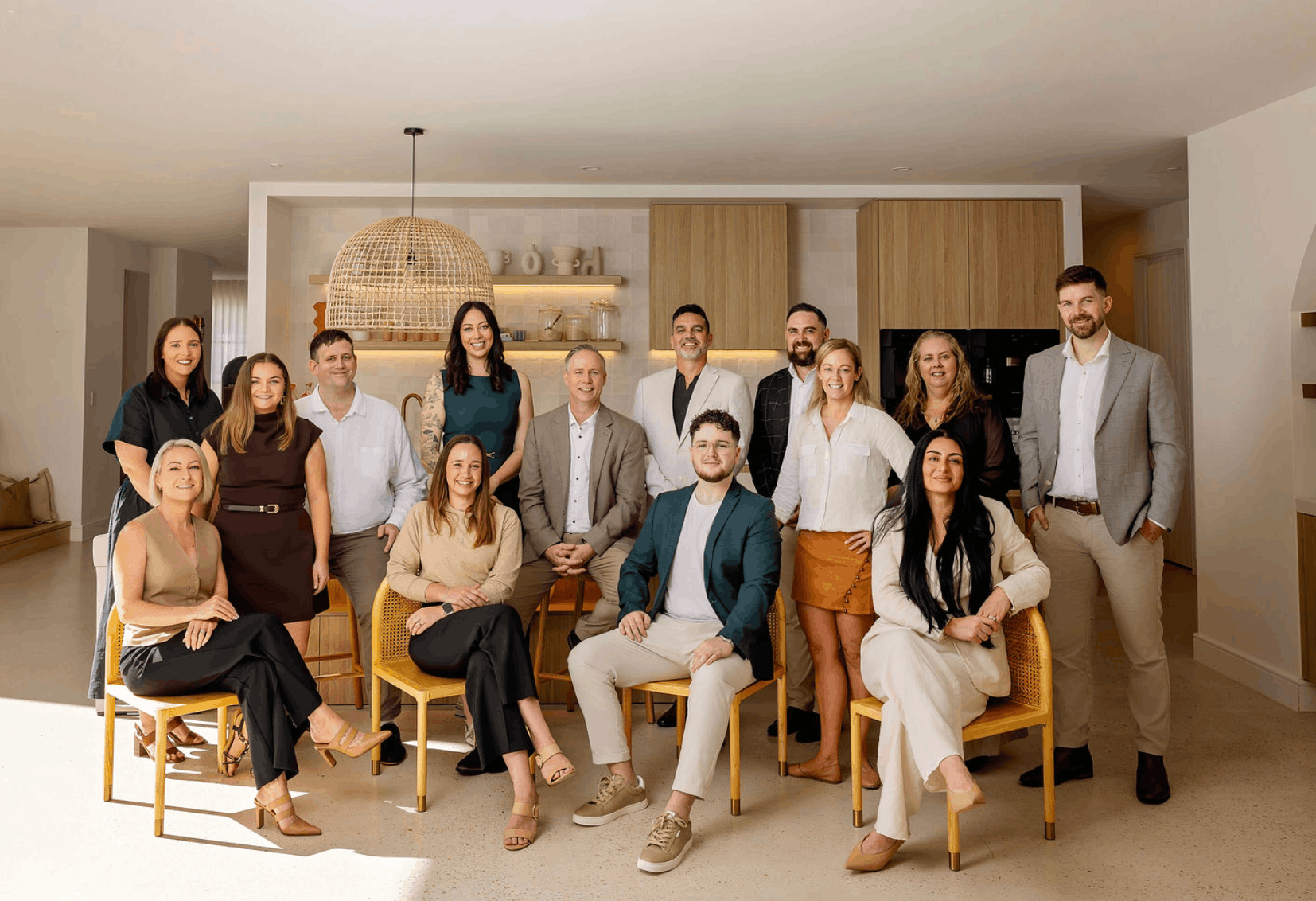 Group of people posing in a bright room with wooden chairs and shelves, likely a team or family portrait.