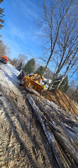 A yellow heavy construction vehicle sits in a snowy, wooded area next to a pile of dirt and logs.