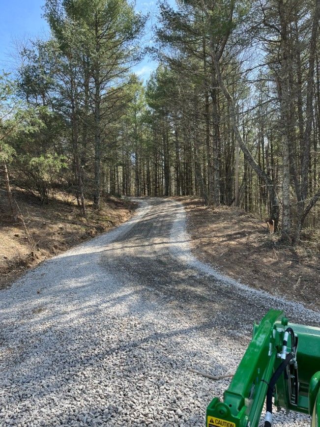 Gravel driveway through a forest of tall trees. A green tractor's arm is in the lower right corner.
