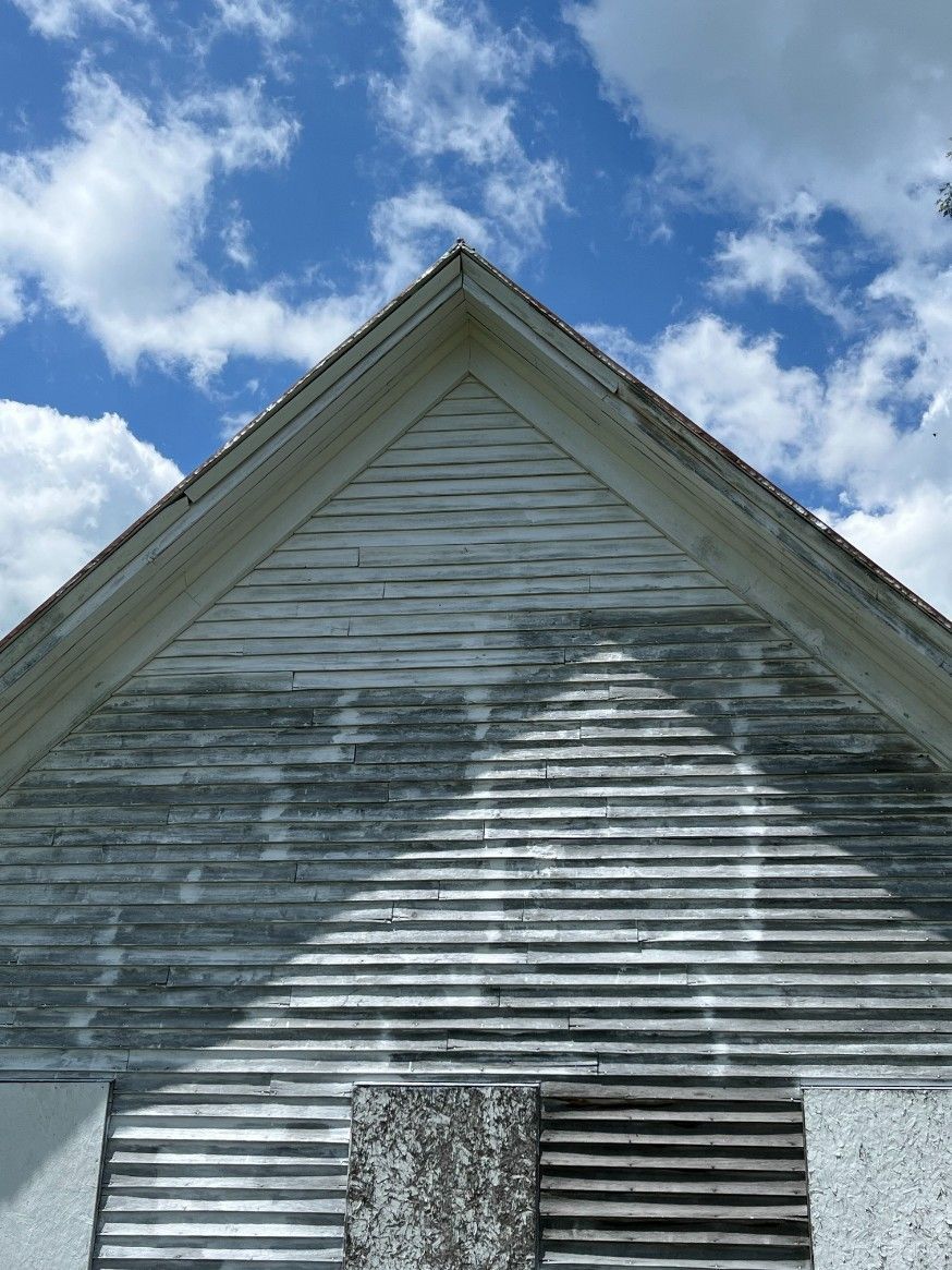 Triangular white building gable against a blue sky with clouds. Boards are weathered and boarded up.