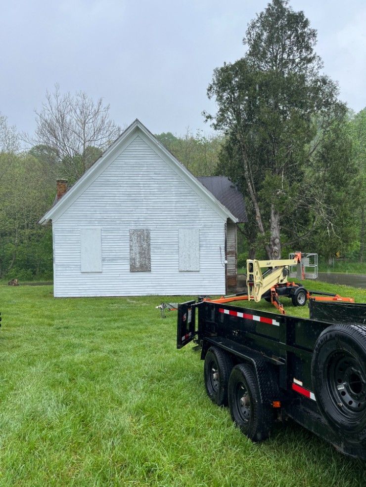 White clapboard building with boarded-up window and trailer on green lawn in front of trees.