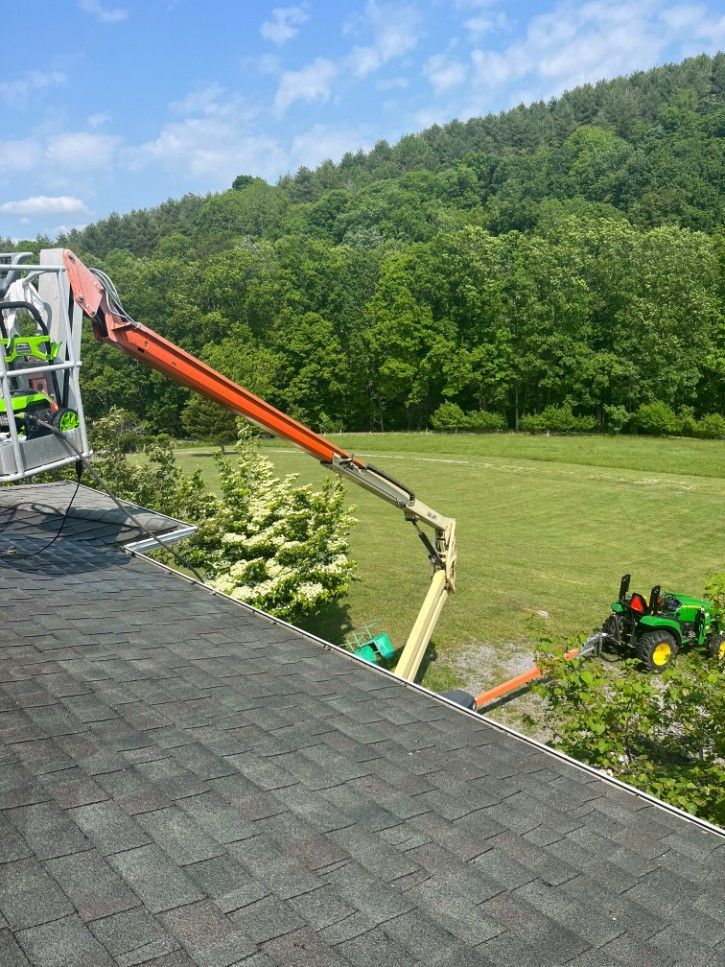 Cherry picker extending from rooftop; a tractor is parked on grass. Green trees and a hillside in background.