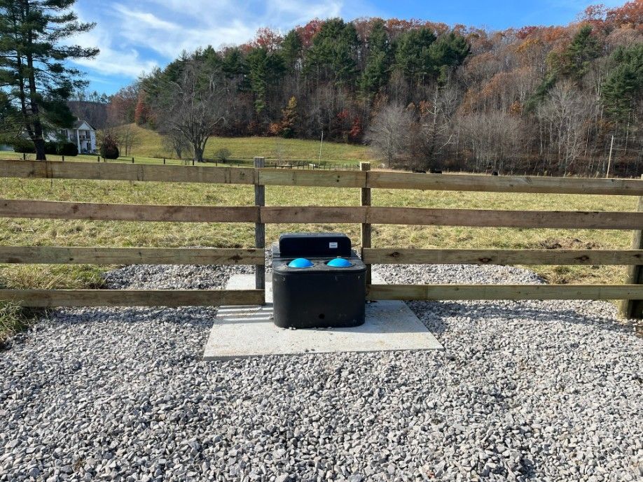 Black water trough in front of a wooden fence with a field and trees in the background.