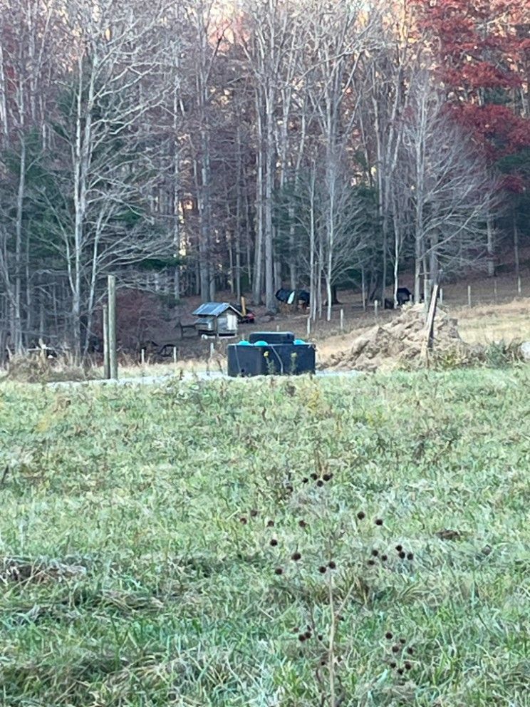 Grassy field, forest background. Large black water tank, small shed, fence, trees.