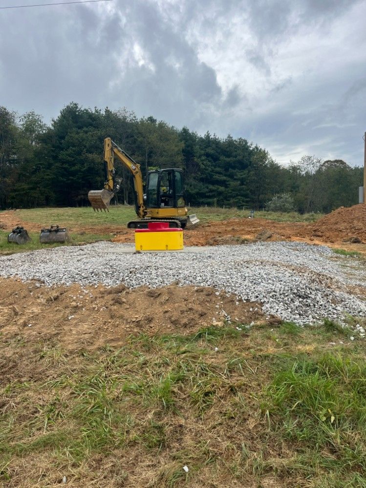 Construction site with excavator and yellow and red box on a gravel bed. Trees in background, overcast sky.