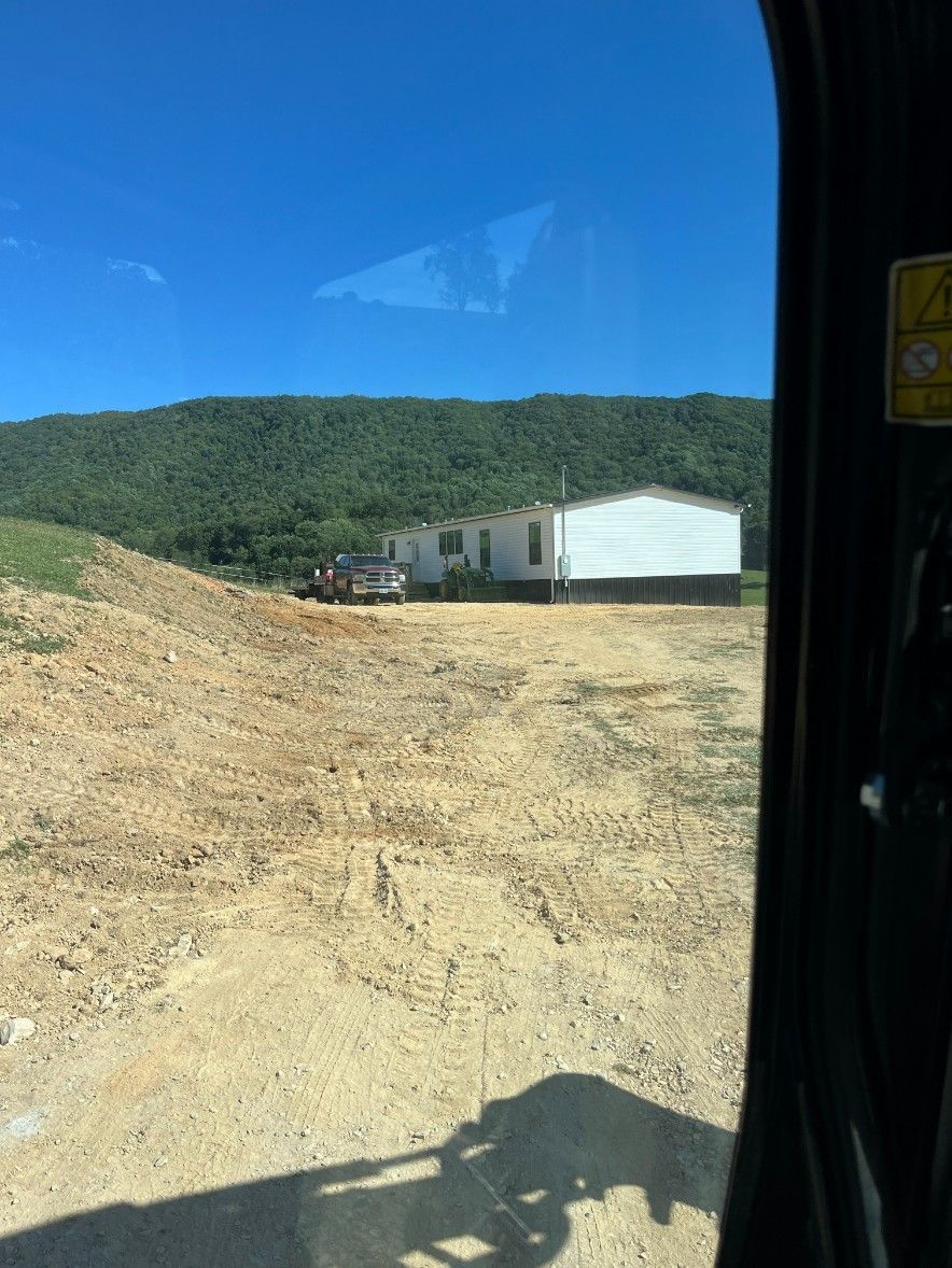 View from machinery cab shows a white house and truck on cleared land with a green mountain backdrop.