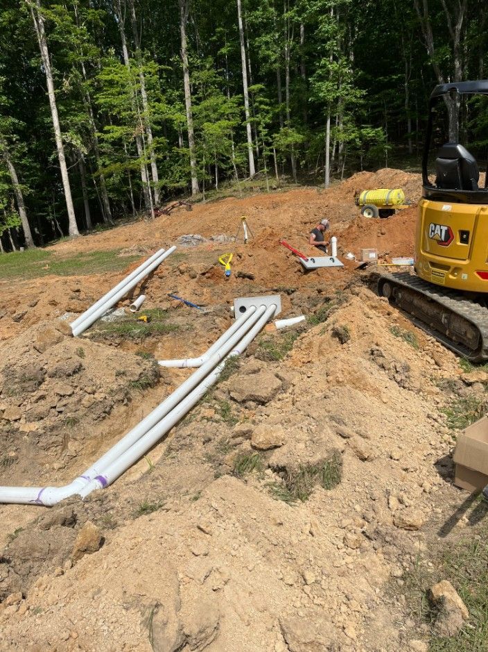 Construction site with trenches, PVC pipes, a backhoe, and equipment. A wooded hillside is in the background.