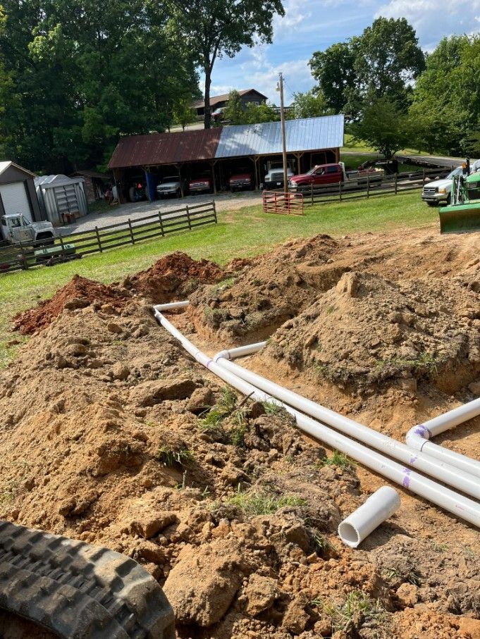 White pipes laid in dirt trenches in front of a barn.