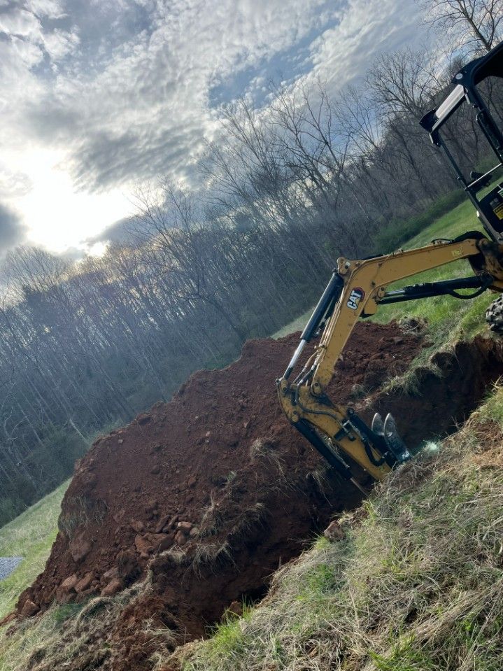 Yellow excavator digging trench in brown earth, grass, and trees under cloudy sky.