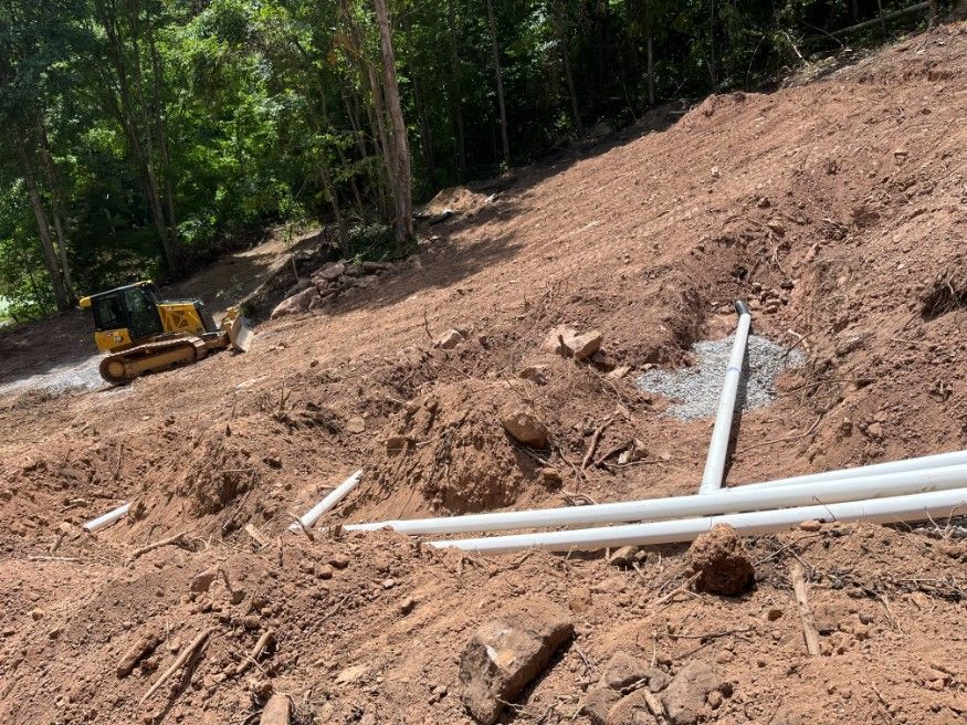 Construction site: excavator on a hillside, white pipes, exposed soil and trees in background.