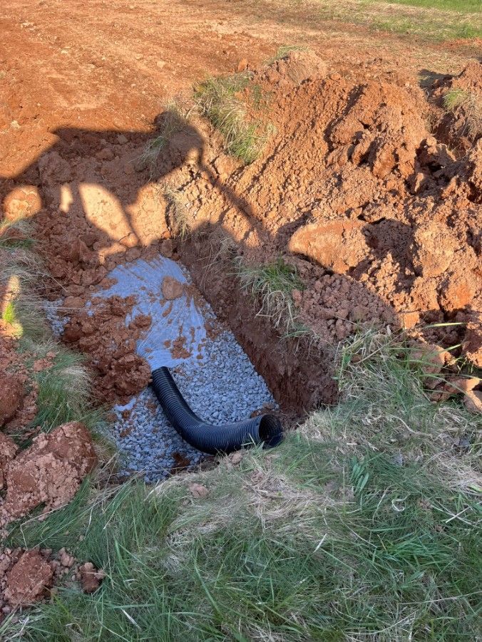 Black pipe emerging from a gravel-filled trench in a grassy area, surrounded by dirt.