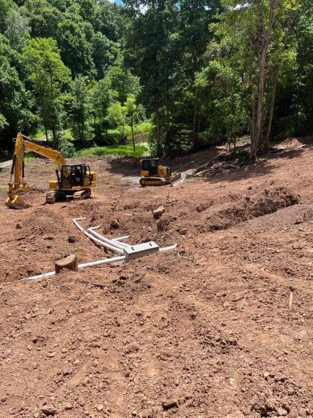 Construction site with two yellow excavators on a dirt lot. White pipes are laid in a trench. Trees in the background.