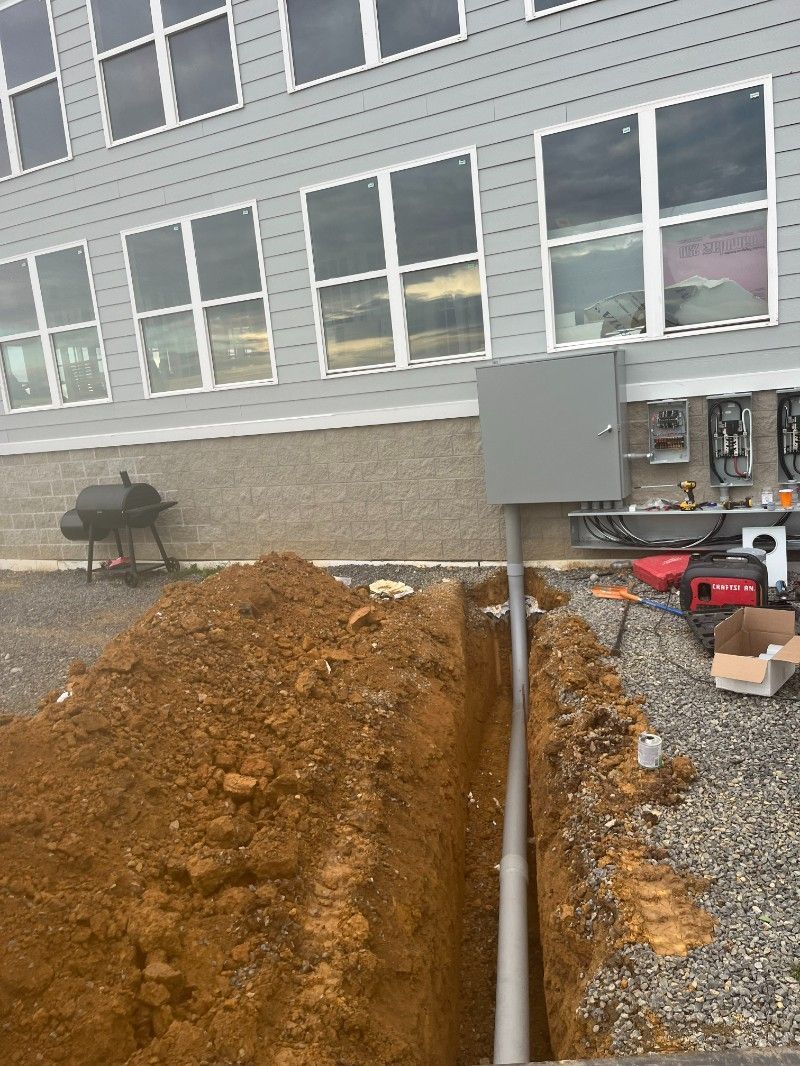Trench dug beside a building. Conduit runs from electrical box to ground. Brown dirt and gravel visible.