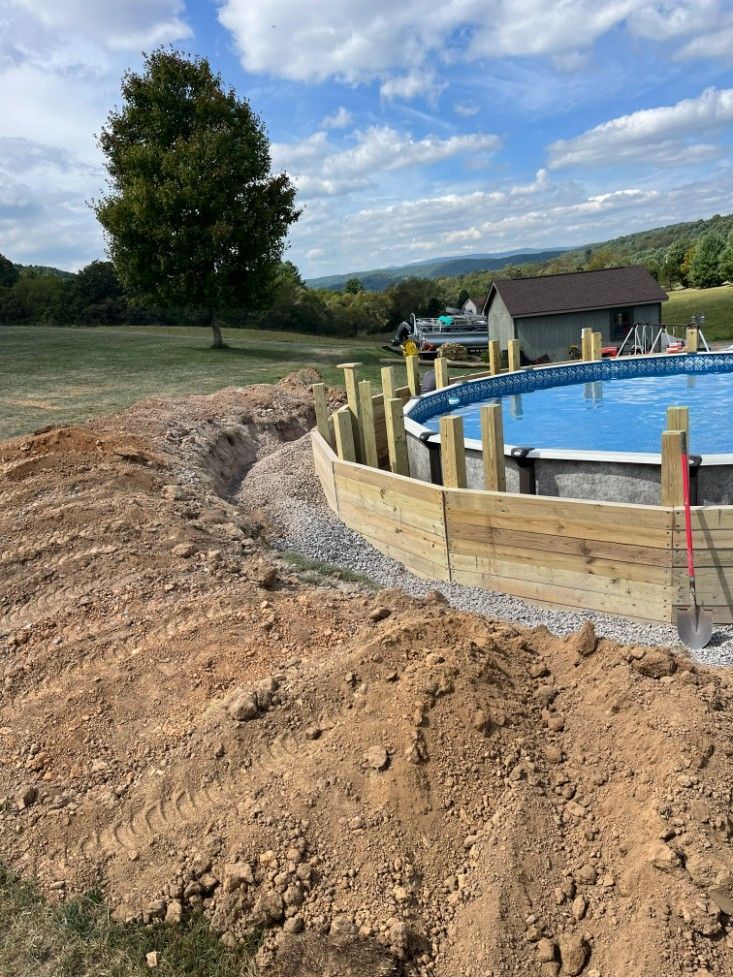 A partially constructed above-ground pool with a surrounding retaining wall on a grassy hillside under a partly cloudy sky.