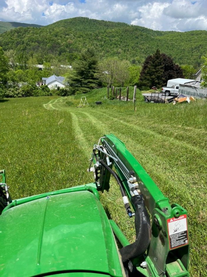 A green tractor mowing a grassy field with a mountain backdrop under a cloudy sky.