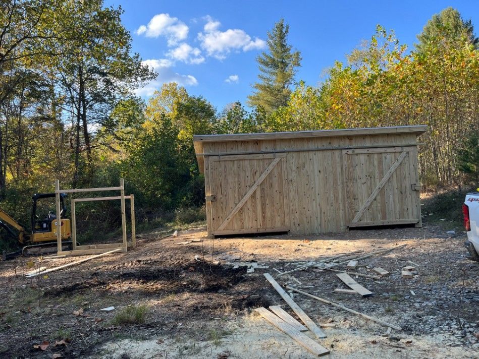 Wooden shed under construction in wooded area, with a small excavator nearby.