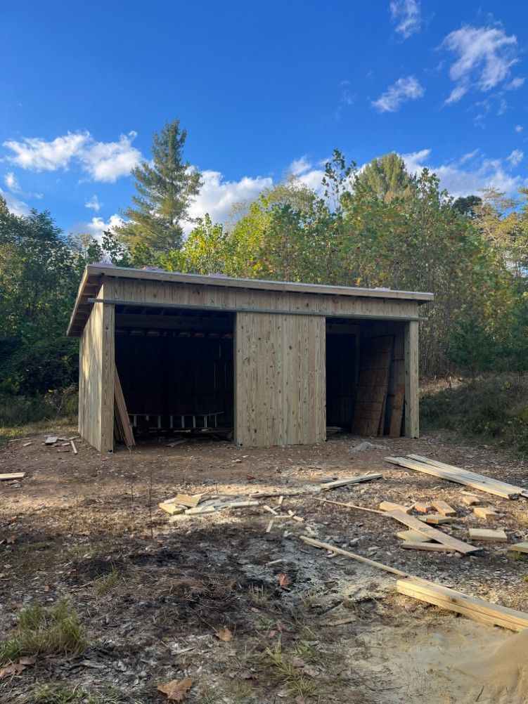 Wooden shed under construction, standing in a clearing amidst trees and a blue sky with clouds.