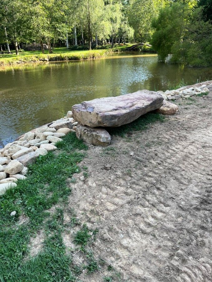 Stone bench by a pond, green trees in background.