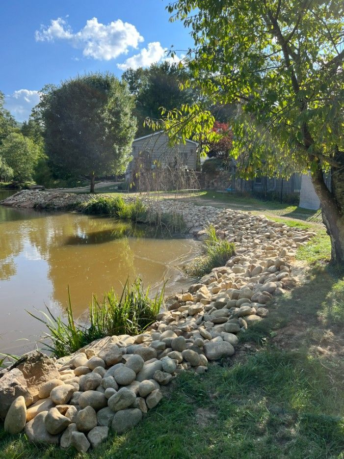Pond with rocky shoreline, lush green grass, and stone building in the background. Sunny day with blue sky.