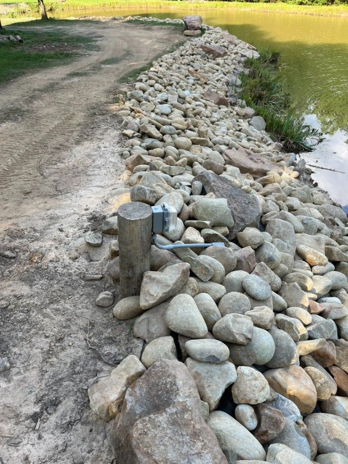 Rocks lining the edge of a pond, next to a dirt path and electrical box.