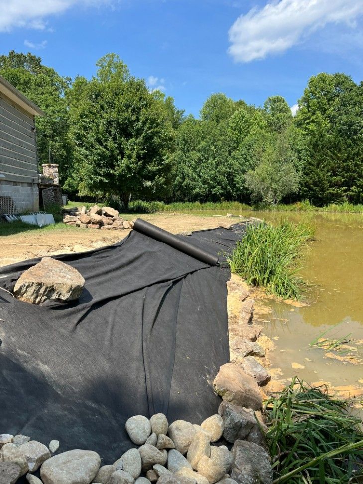 Black landscape fabric on pond edge, rocks, tall grass, green trees, sunny day.