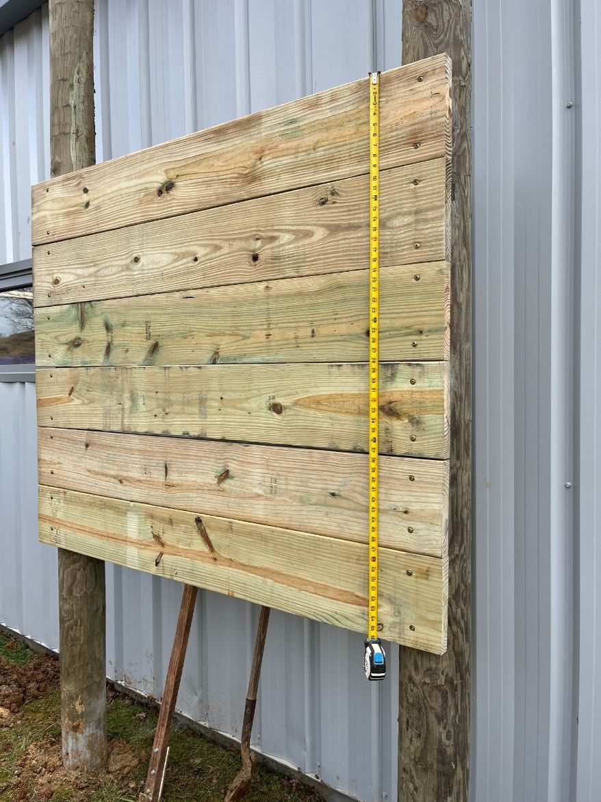 Wooden target with a yellow tape measure, attached to a metal post in front of a metal building.