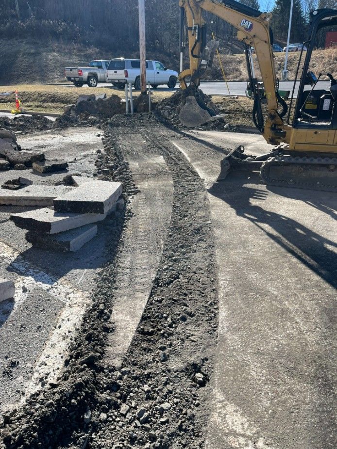 Excavator digging a trench in pavement. Concrete slabs and a truck are visible in the background.