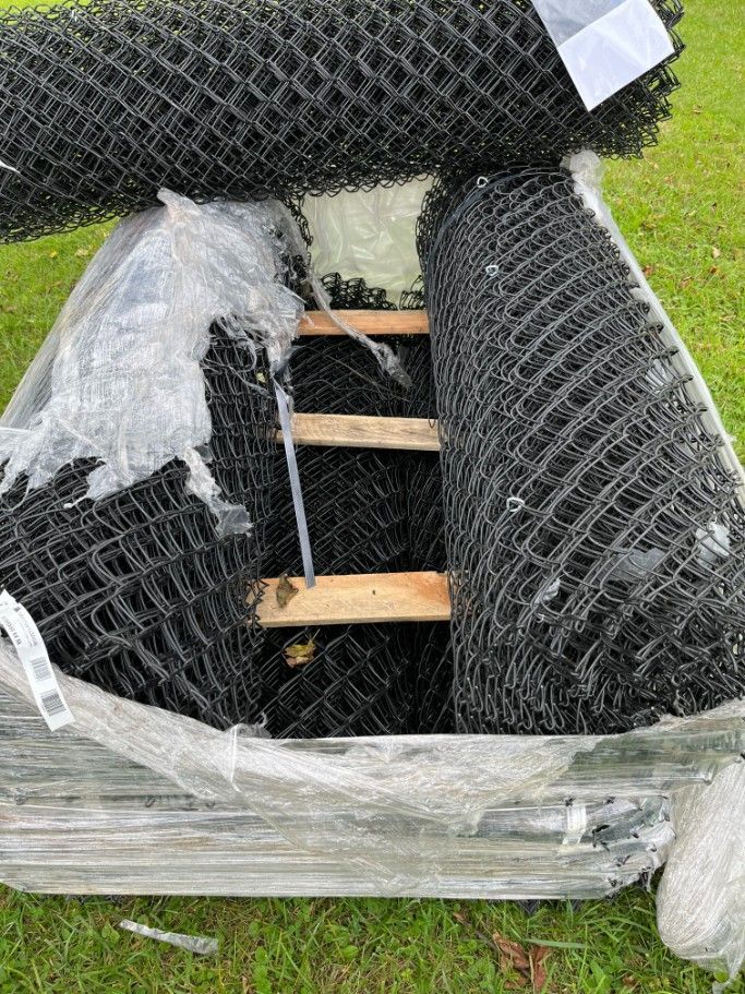 Black chain-link fence rolls on a wooden pallet, wrapped in clear plastic, outdoors on grass.