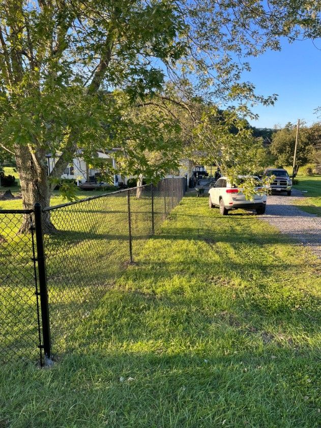 Chain-link fence in front of a house, a white SUV, and a driveway with another vehicle. Sunlight on the grass.
